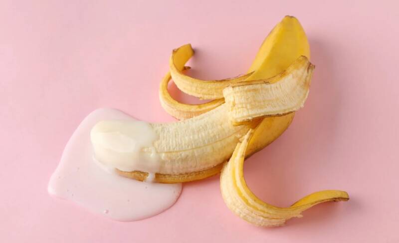 A peeled banana on a pink surface with a creamy white substance on it, suggesting a suggestive visual metaphor